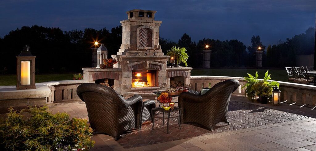 A grand outdoor fireplace at night, built with Unilock stone pavers and a tall chimney. The fireplace has a lit firebox and is surrounded by comfortable wicker chairs on a brick-style paver patio.