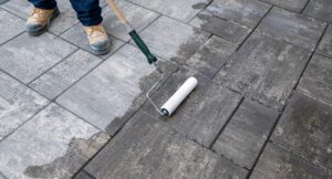 A professional using a paint roller to apply sealant to a Unilock paver patio, with one half of the patio appearing damp and darker from the applied sealant.